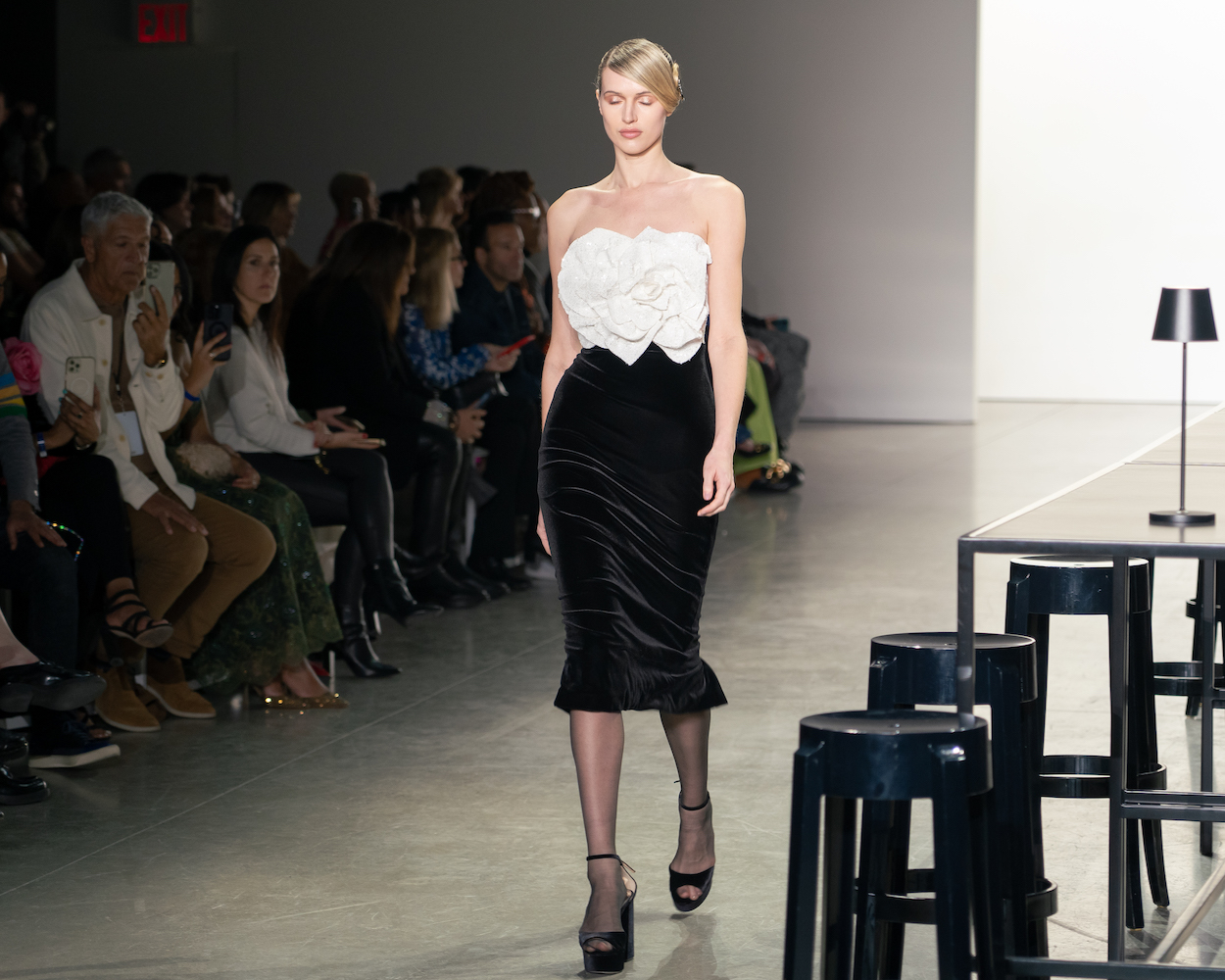 A model walking down the runway wearing a white, rose-shaped, ruffled top, a black velvet skirt and black heels.