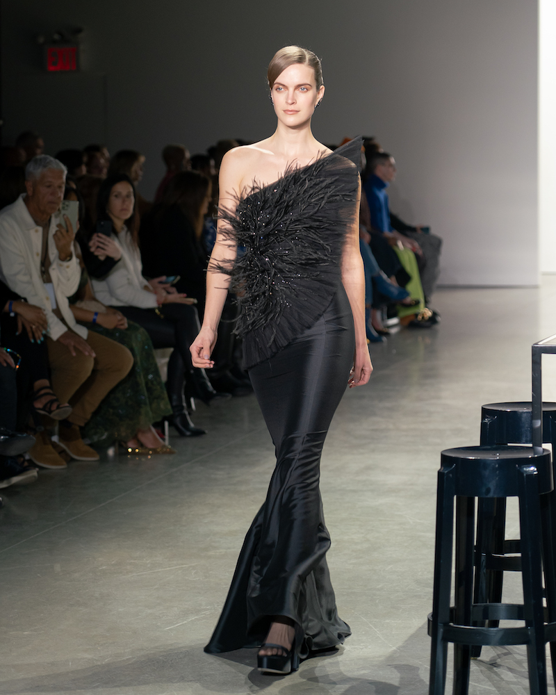 A model walking down the runway wearing a black feathery top, a long black silky skirt and black heels.