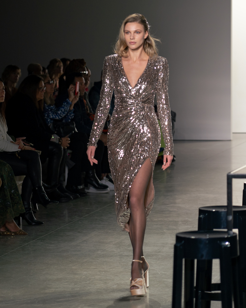 A model walking down the runway wearing a sparkly gold long-sleeved mini dress and champagne heels.
