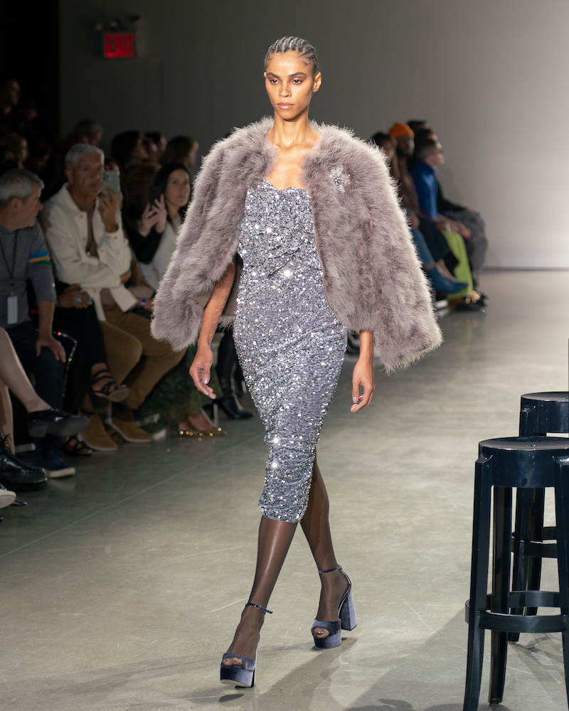 A model walking down the runway wearing a sparkly pastel purple mini dress, a purple fur coat and navy heels.