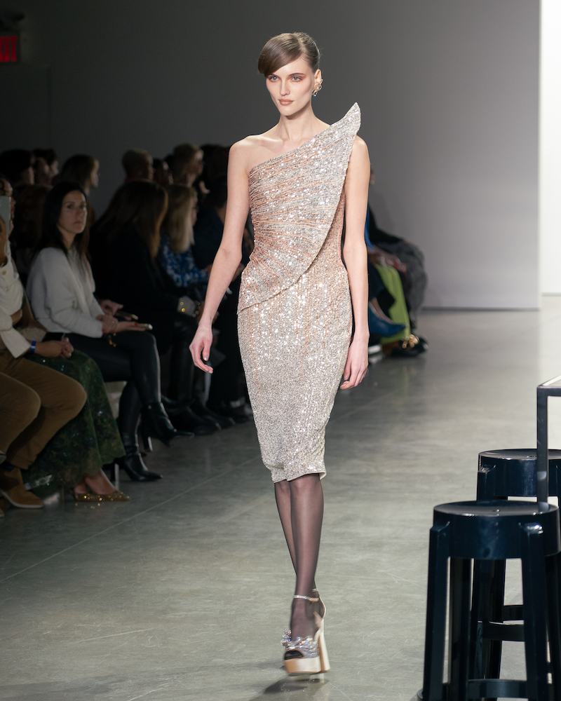 A model walking down the runway wearing a geometric sparkly silver mini dress and champagne heels.