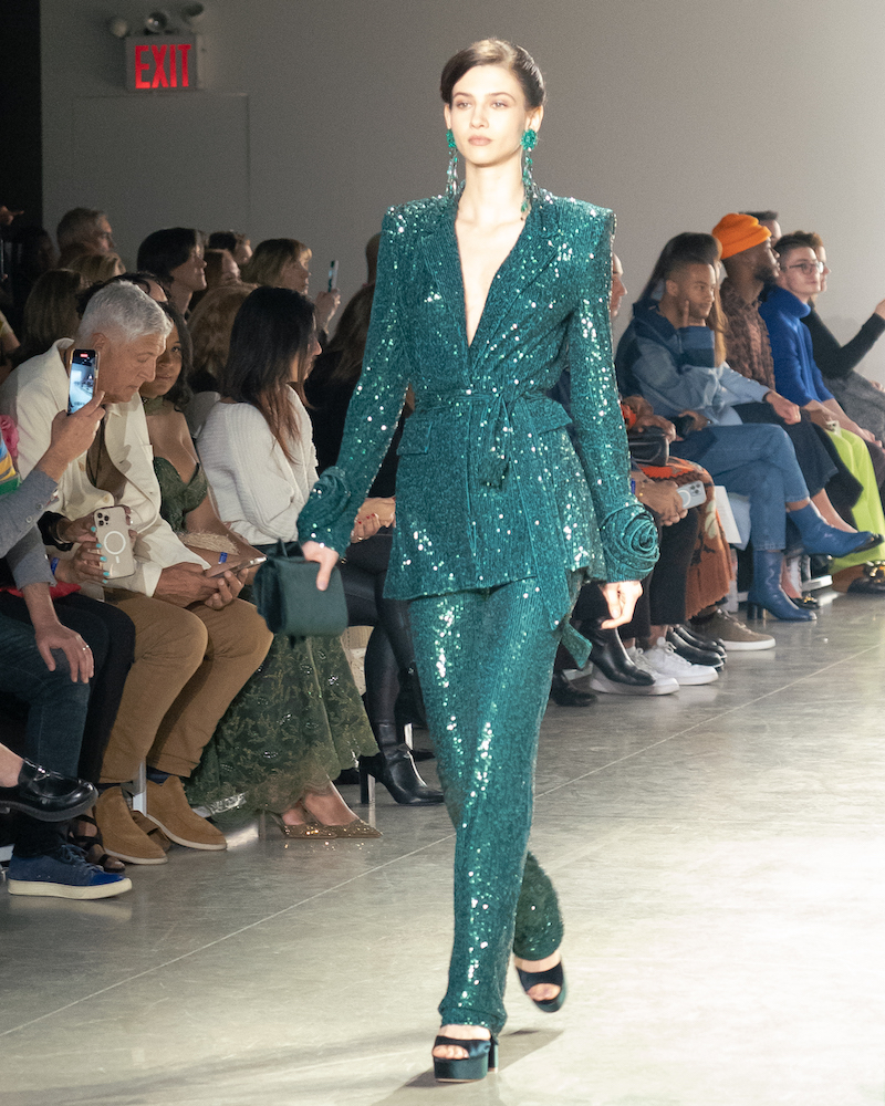 A model walking down the runway wearing a sparkly, teal matching pant and blazer set with sparkly teal earrings and teal heels.