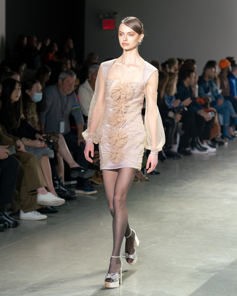 A model walking down the runway wearing a lacy, pastel cream, long-sleeved mini dress with champagne heels.