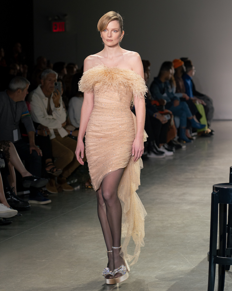 A model walking down the runway wearing a pastel cream mini dress with a feather-lined top and champagne heels.