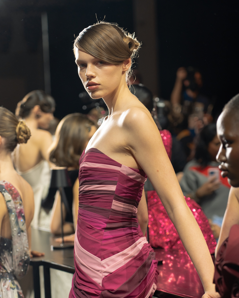 A model posing on the runway wearing a pink, maroon and red mini dress, looking into the camera, surrounded by other models at the end of the show.