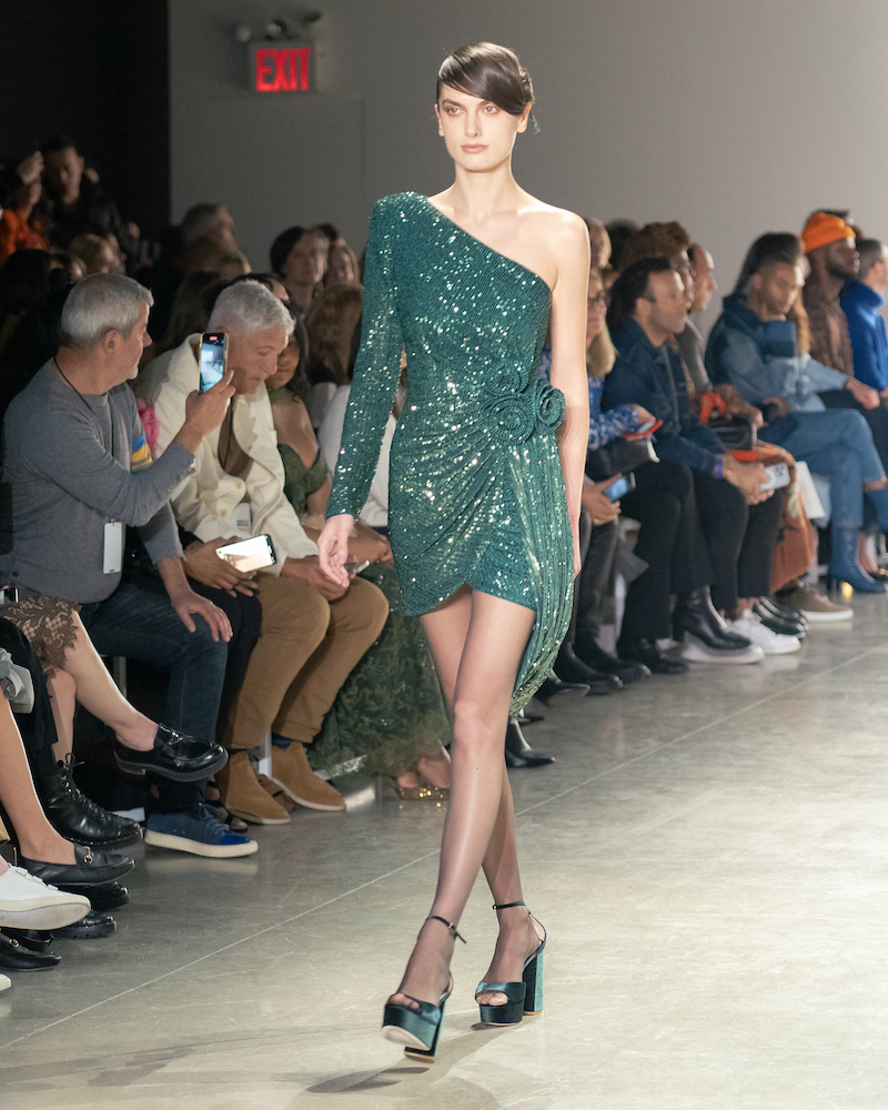 A model walking down the runway wearing a sparkly, teal dress with one sleeve and teal heels.