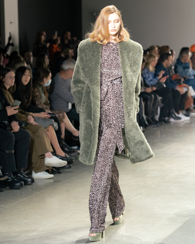 A model walking down the runway wearing a sparkly matching pant and top set, a long green fur coat, and matching green heels.
