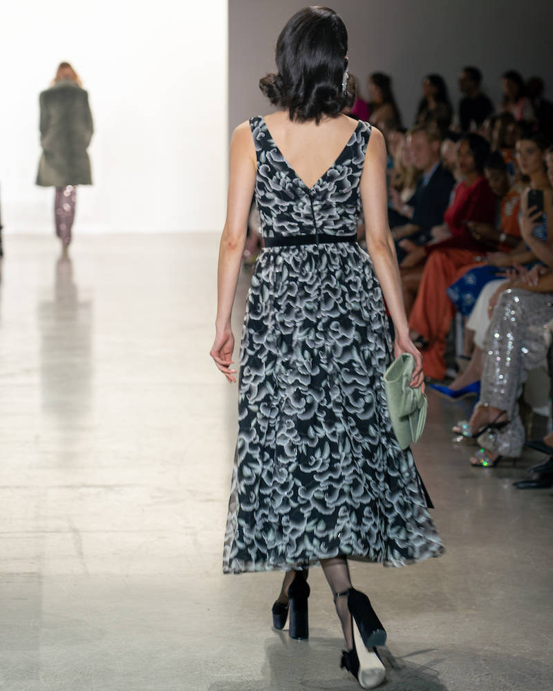A model walking down the runway, pictured from the back, wearing a patterned blue and black dress, black heels and a green bag.