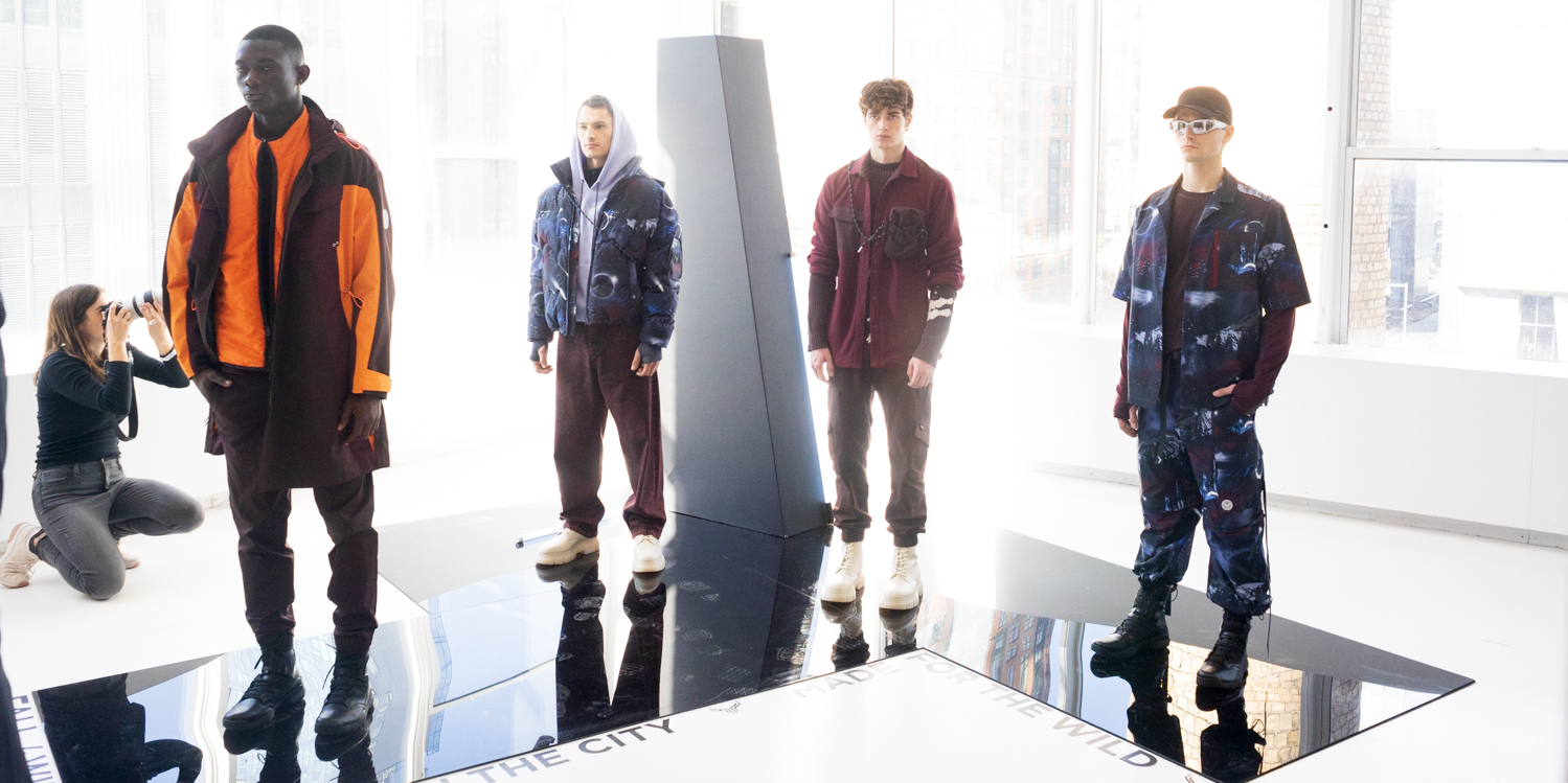 Four models stand in a line in a white room with large windows in the back. The model on the far left is wearing a black and orange jacket, black pants and black boots. The second model on the left wears a gray hoodie, blue and black puffer jacket, brown pants and white shoes. The model on the far right wears a long-sleeve maroon shirt, a patterned button-up shirt and pants, black boots, white sunglasses and a black baseball cap. The second model on the right wears a black, long-sleeved shirt, a maroon button-up shirt on top, brown pants and white boots.