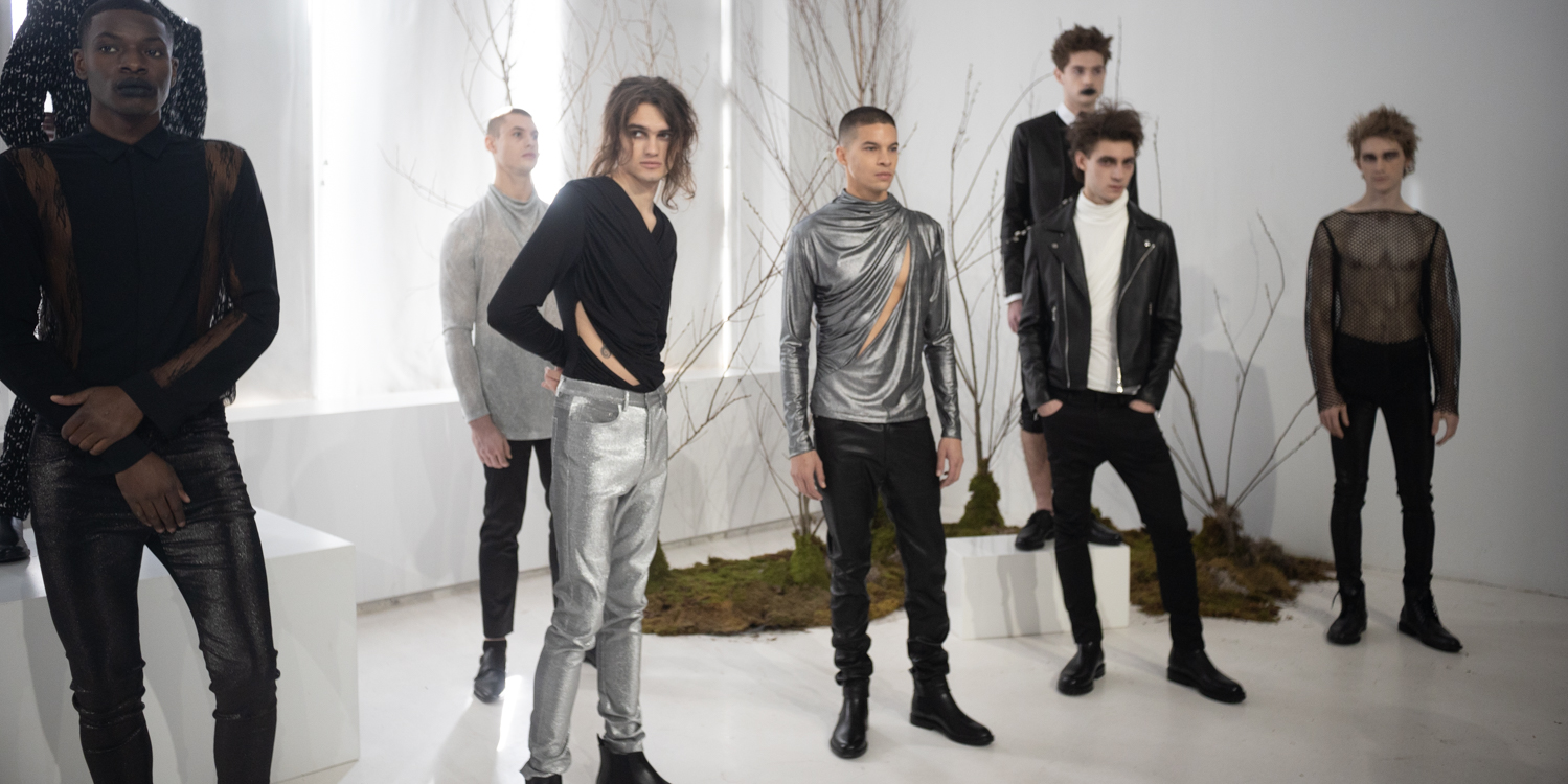 A group of eight models stand wearing various black, white and silver outfits. Two models on either side of the room stand on white boxes. Behind them are four leafless trees.