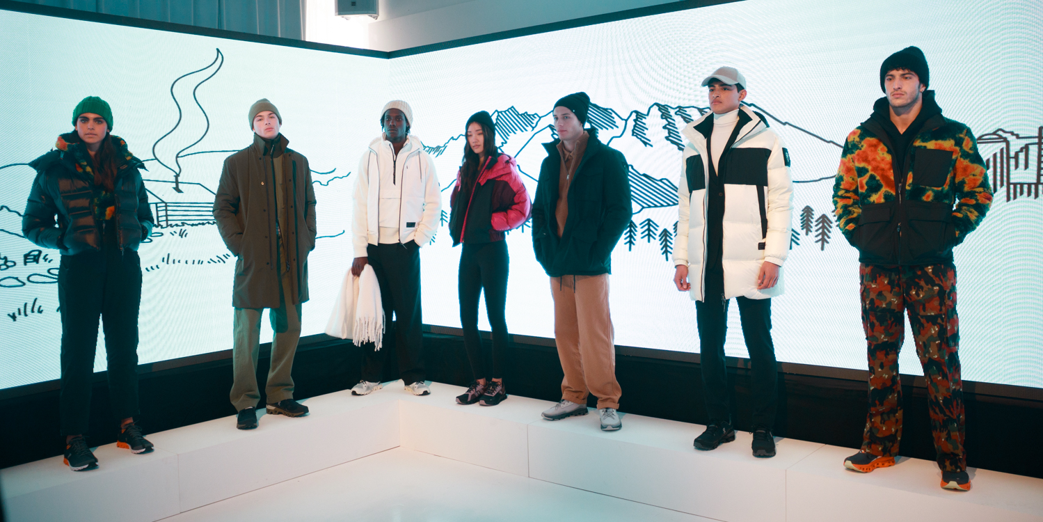 Seven models stand in a line, each wearing jackets, pants and beanies in various colors. Behind them is an LED display with sketches of a ski-lift and trees.