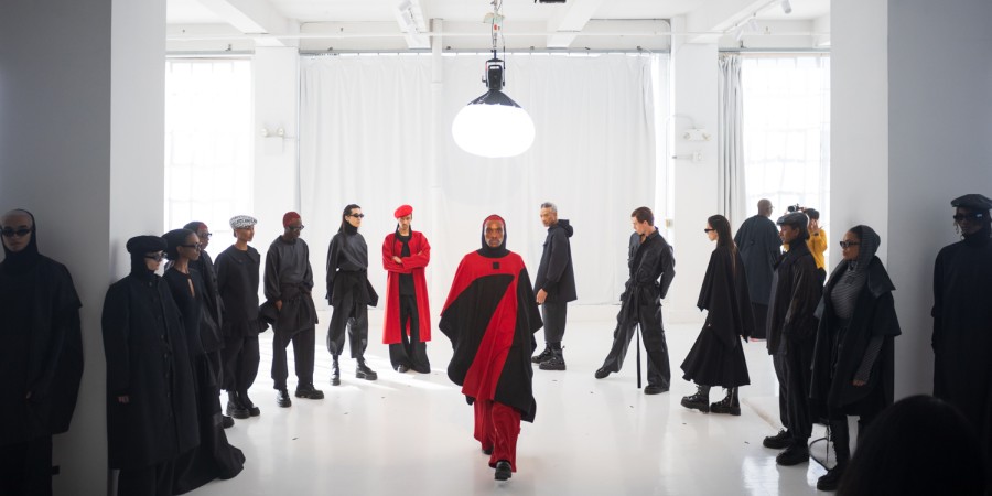 A group of models stand in a semi-circle formation wearing black and red outfits. One model walks from the circle into the middle of the room. Above him is a large, round light fixture hanging from the ceiling.
