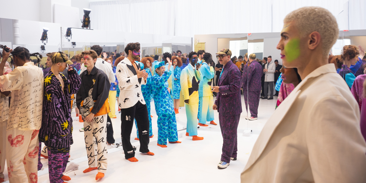 A group of models is pictured standing in front of a large mirror. The models are dressed in clownlike outfits, and the designer, dressed in a patterned purple suit, stands in the middle, directing the models.