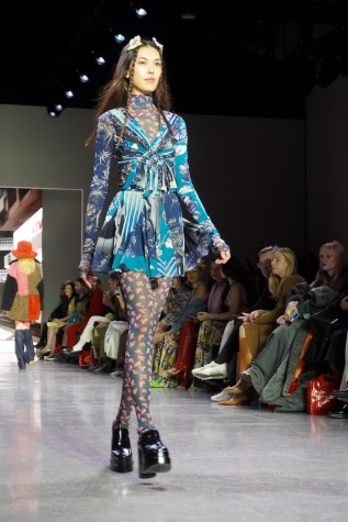 A model walks down the runway wearing N.F.T. hair accessories, a blue patterned minidress, leggings with Zodiac sign prints and black boots.