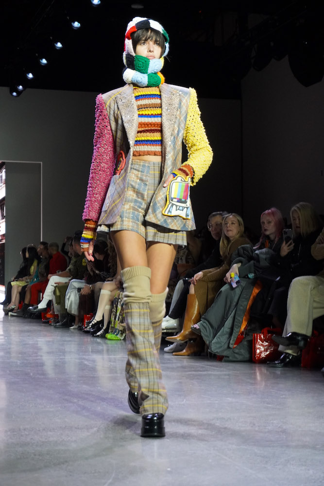 A model walks down the runway wearing a striped sweater, a plaid blazer, a plaid skirt, plaid leg warmers, a colorful balaclava and black boots.