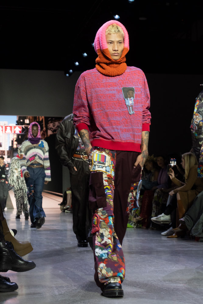 A model walks down the runway wearing a coral and lavender sweater with an N.F.T. drawing, cargo pants, a balaclava and black boots.
