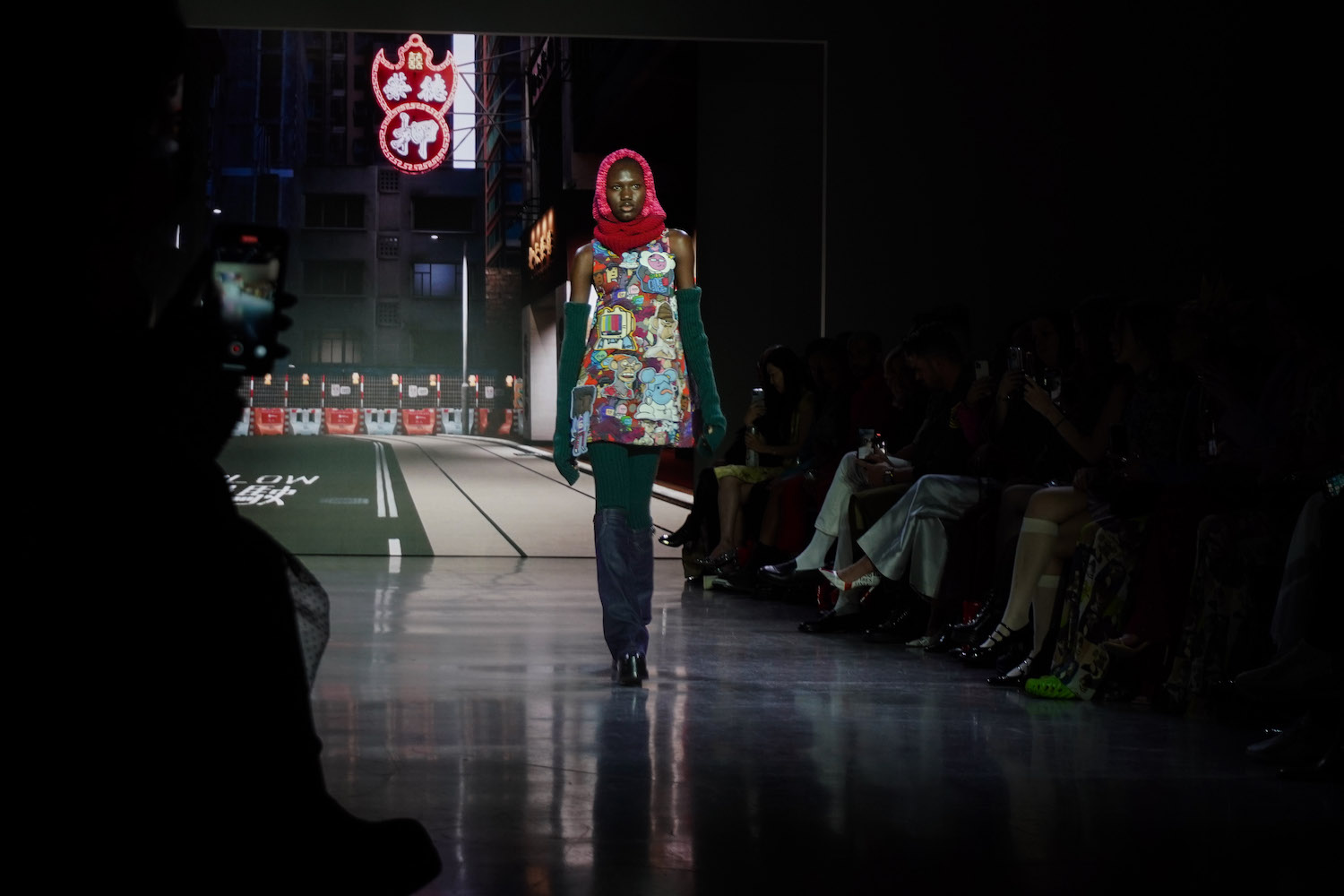 The opening model of the show walks down the runway in dim lighting against the TAMverse backdrop with a dress decorated with N.F.T.s, green leggings and arm warmers, pink and red sleeves, and tall boots.