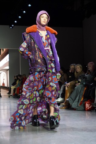 A model walks down the runway wearing a long, ruffled purple dress with Zodiac sign designs, a sequin jacket, a lavender balaclava and black boots.