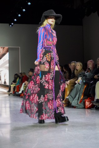 A model walks down the runway wearing a blue and pink striped long-sleeve top, a decorated mesh dress with a slit down the middle, a black fluffy hat, tall black leg warmers and black boots.