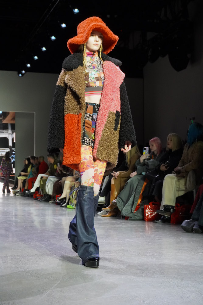 A model walks down the runway wearing an orange, fluffy hat, a patterned crop top, a patterned skirt, flower-patterned leggings, a fleece coat, tall black leg warmers and black boots.