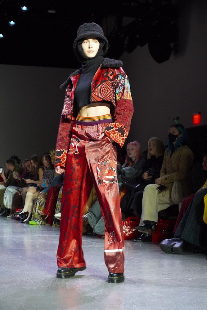 A model poses on the runway wearing a black turtleneck crop top, a red jacket, red pants, a black fleece hat and black boots.