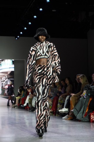 A model walks down the runway wearing a black fleece hat, a black-and-white striped crop top, jacket and pants, along with black boots.