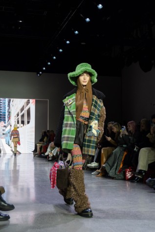 A model walks down the runway wearing a green plaid jacket, a brown balaclava, a green hat, a patterned top and leggings, a leather skirt with a belt, brown leg warmers, black boots and a bag.