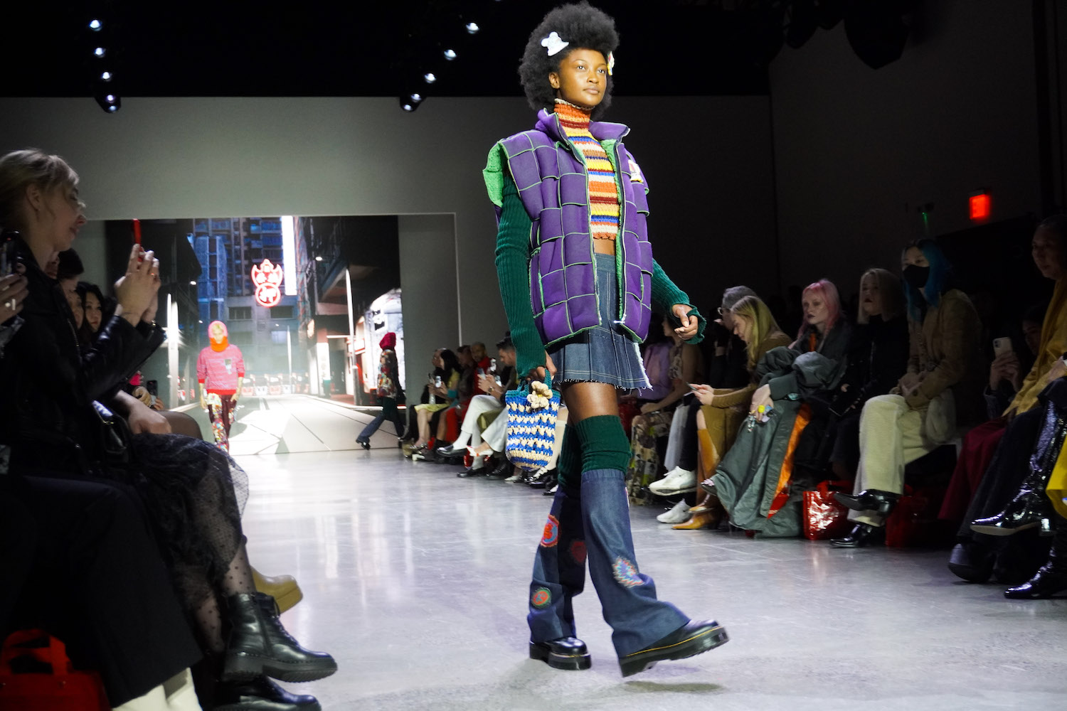 A model walks down the runway wearing a striped sweater, a purple woven jacket with green sleeves, high green socks, denim leg warmers, a blue-and-white crocheted bag and black boots.