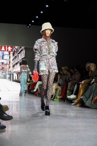 A model walks down the runway wearing an N.F.T.-patterned jacket and jeans, a zodiac sign-patterned top and leggings, a brown balaclava and black boots.