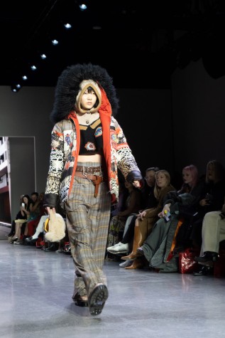 A model walks down the runway wearing a black top, a patterned coat with a fluffy hood, plaid pants, two brown belts and black boots.