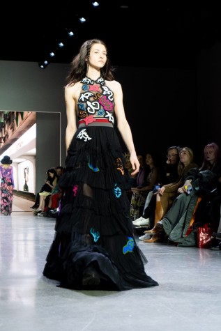 A model walks down the runway wearing an N.F.T.-patterned high-neck top, a black ruffled skirt and black boots.