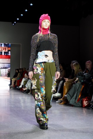 A model walks down the runway wearing a pink balaclava, a sparkly crop top, green patterned pants and black boots.