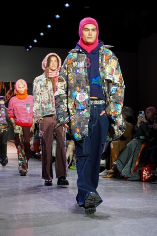 A model walks down the runway in the final runway look, wearing a pink balaclava, a navy blue sweatshirt, N.F.T.-patterned outerwear, jeans and black boots. Two models walk behind them.