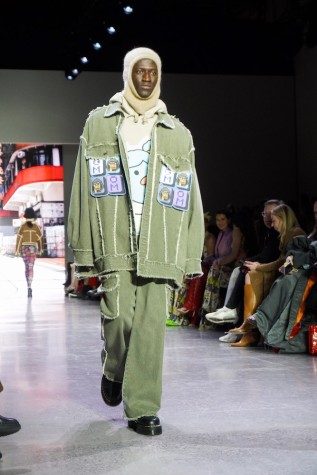 A model walks down the runway wearing a beige balaclava, a beige sweater, a green jacket and pants, along with black boots.