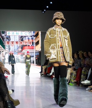 A model walks down the runway with a brown woven fleece hat, a patterned top and skirt set, a beige jacket stitched with a Bathing Ape N.F.T. and white borders, high black socks, green leg warmers and black boots.