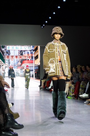 A model walks down the runway with a brown woven fleece hat, a patterned top and skirt set, a beige jacket stitched with a Bathing Ape N.F.T. and white borders, high black socks, green leg warmers and black boots.