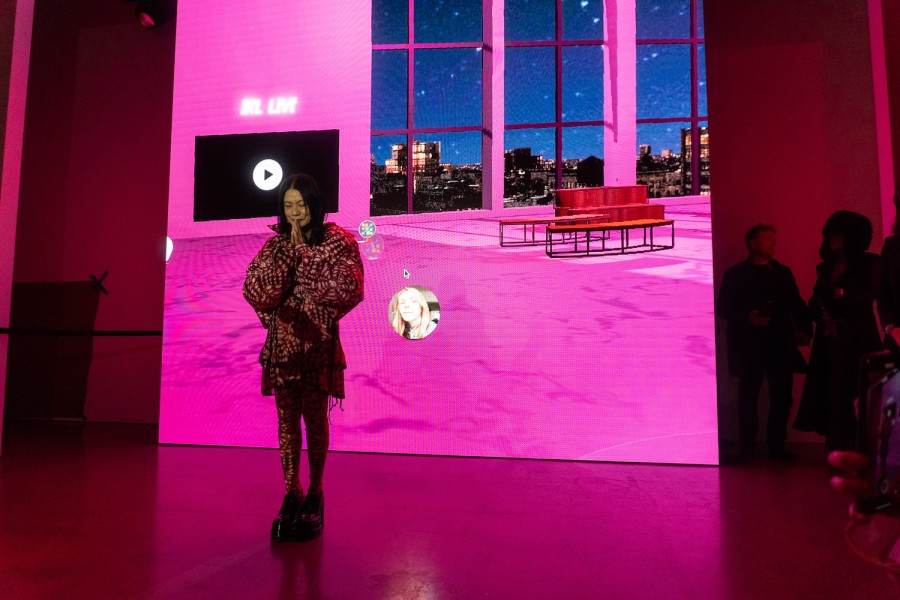 Vivienne Tams wears a red and white patterned dress. She prays with two hands enclosed in front of her mouth, standing in front of a purple backdrp.