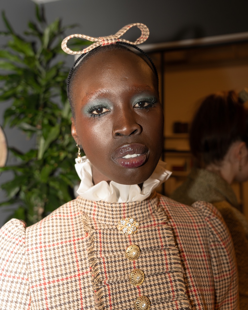 A photograph of a model looking into the camera, wearing blue eyeshadow, an amber tweed jacket with a ruffled white collar and chunky buttons, and a matching bow.