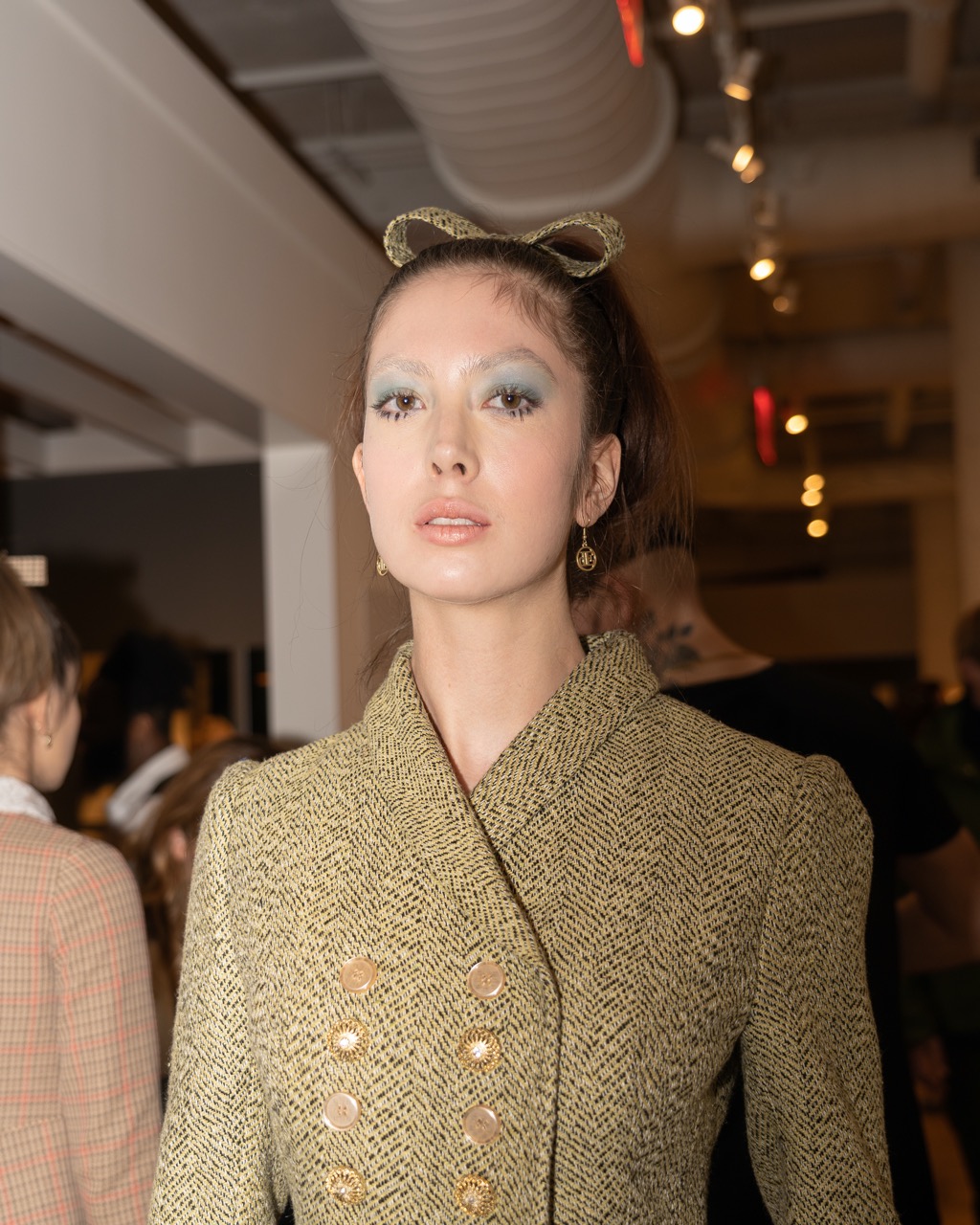 A photograph of a model looking into the camera, wearing blue eyeshadow, a muted yellow tweed jacket, and a matching bow on her head.