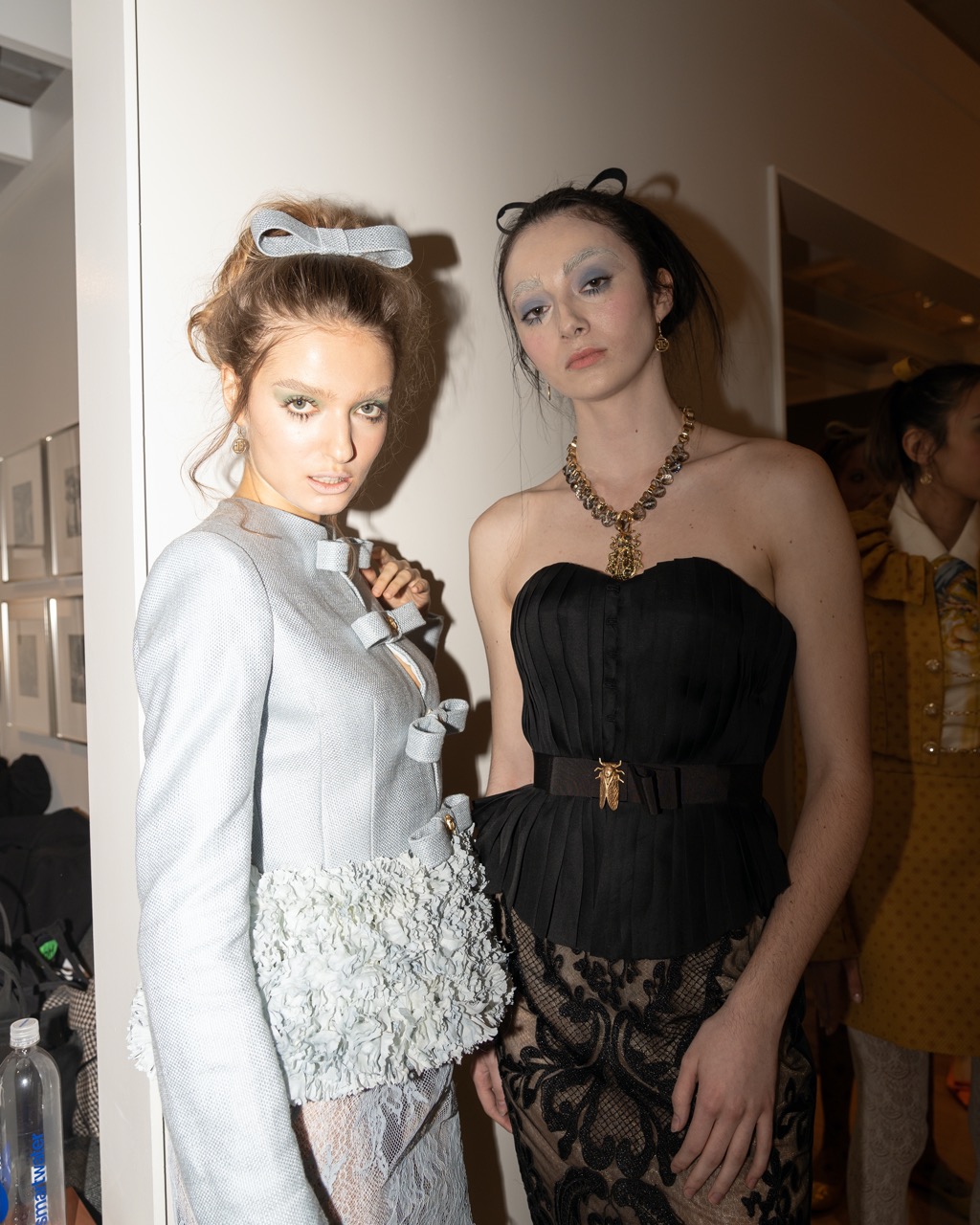 A photograph of two models looking into the camera, wearing blue eyeshadow. The model on the left wears a dusty blue set lined with bows that match the one on her head. The model on the right wears a black top with a gold insect belt, black mesh pants and a chunky necklace that matches the belt.
