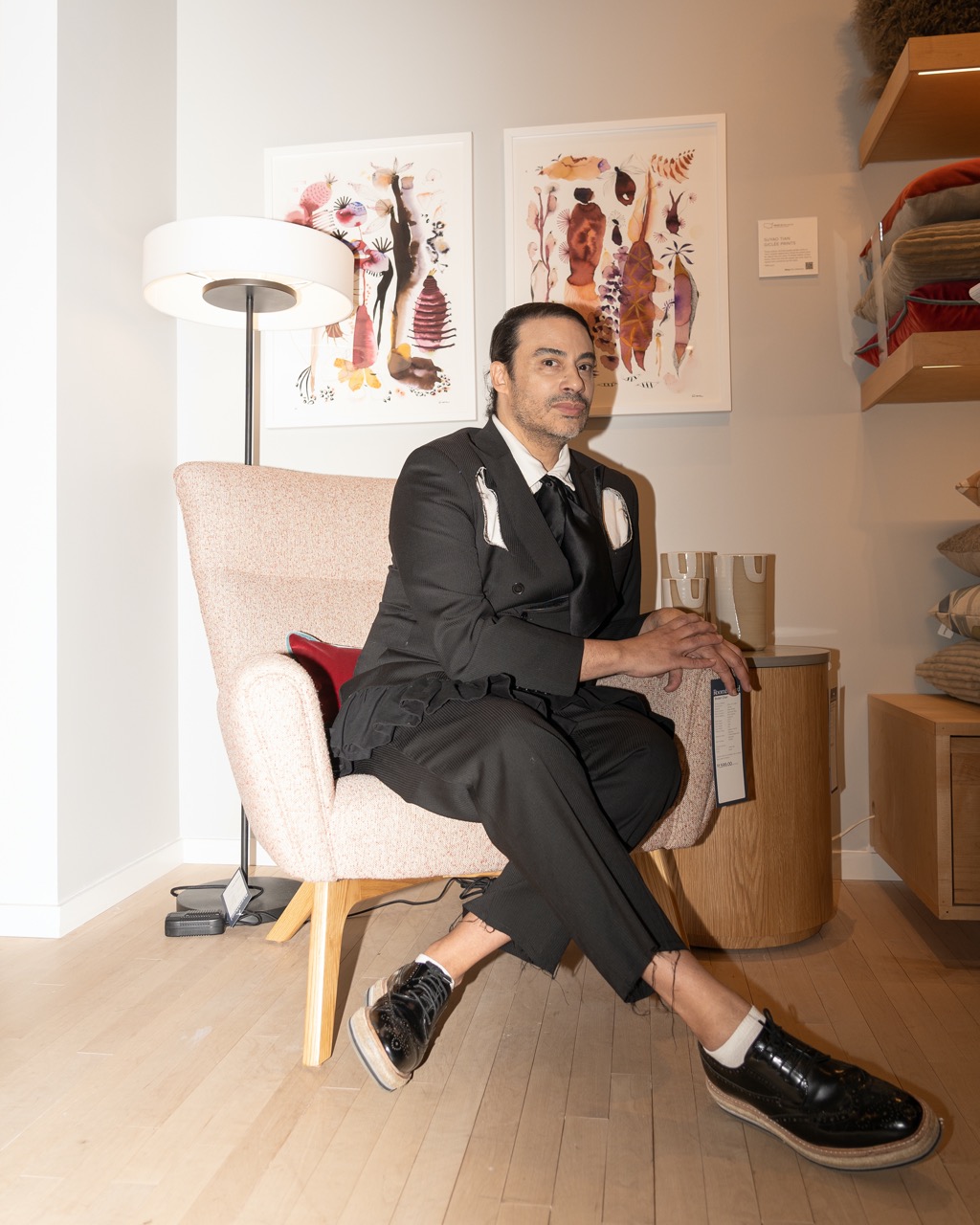 A photograph of Victor, the designer of the collection, sitting in a chair and looking into the camera. He is wearing a black suit and black shoes.
