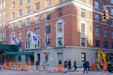 The exterior of NYU’s Rubin Hall on Fifth Avenue. There are two NYU flags hanging outside the entrance with a green sunshade, and there are pedestrians passing by the building. The sun shines on the upper floors of the building.