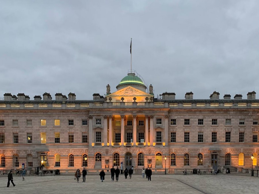 The London Museum beneath a cloudy sky. Beams of orange lights hit the beige brick walls and windows. At the center of the image, a light green dome sits on top of the building and its pillars.