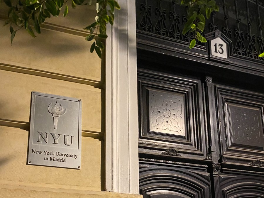 This photo is a tight shot of the exterior of N.Y.U. Madrid’s building. With several green twigs hanging down, the black doors are closed with the number 13 written on the center above. A gray metal sign with the N.Y.U. torch logo and the text “New York University in Madrid” is hung on the yellow wall on the left side of the image.
