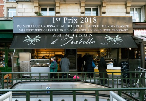 The storefront of a restaurant named “La Maison d’Isabelle” in Paris with white text in French and two light green plants printed on each side of the black sunshade. Several customers are lined up next to the bright yellow counter on the right side of the store, which is warmly and dimly lit.