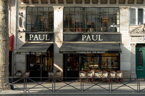 The storefront of a dessert shop named “Paul” in Paris which has many chairs outside. There is white text on the black sunshade that reads “Salon de the” and “Patisserie fine pur beurre.”