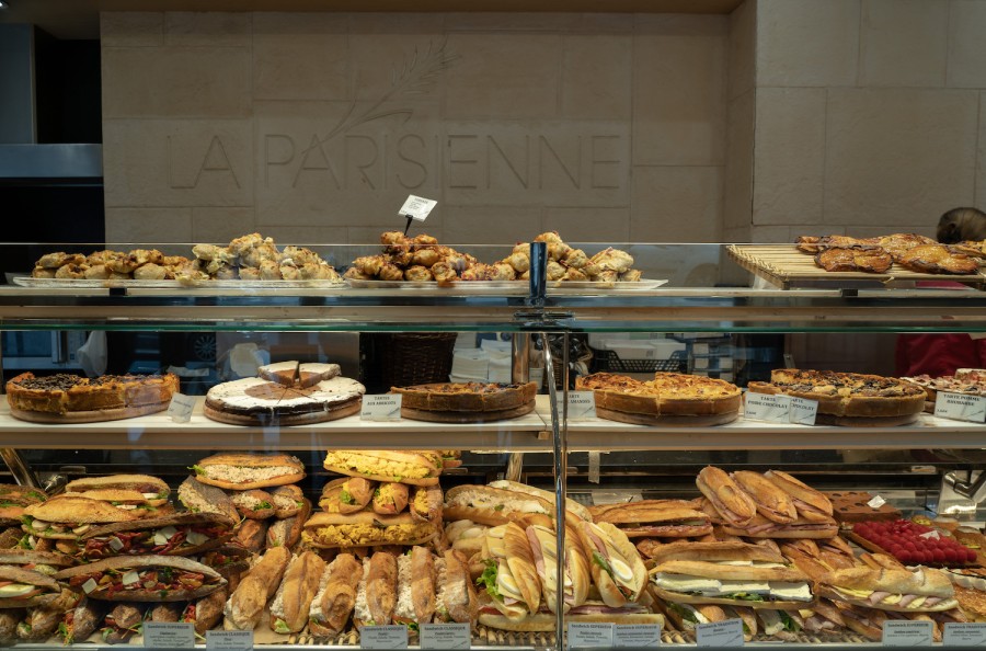 Glass cases with shelves inside a bakery filled with various baguette sandwiches, pastries and pieces of cake. On the white stone brick wall behind the glass cases reads “La Parisienne.”