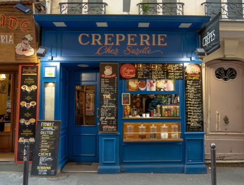 The storefront of dessert shop Crêperie Chez Suzette in Paris with blue paint and pictorial menus on the exterior. A staff member is seen through the cashier window, which is decorated with food pictures above, a soda exhibition on the right corner, a basket of fruit on the left corner and five glass jars of food on the bottom.