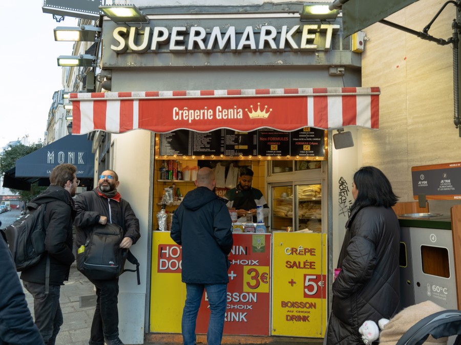 The storefront of Crêperie Genia in Paris with white text that reads “Crêperie Genia” next to a crown printed on its red and white sunshade. White text “Supermarket” is shown above the sunshade. Four customers stand outside of the bright yellow and red counter, and a male-presenting staff is inside next to a glass exhibition with bread shown inside.