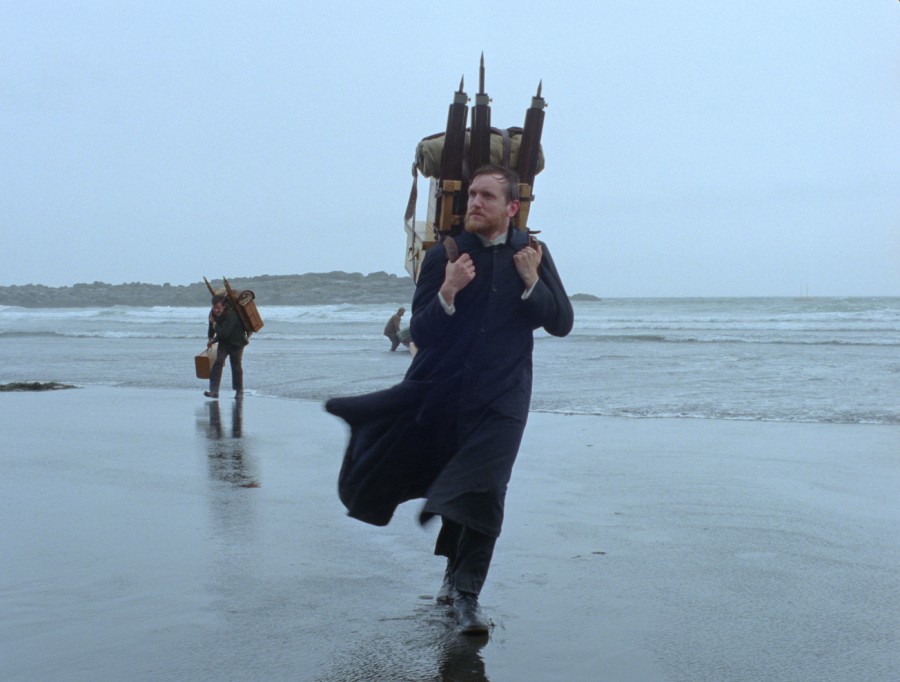 A film still from Godland with actor Elliott Crosset Hove, who plays Lucas, in the center on the beach. The ocean, shoreline, and another character can be seen behind him.